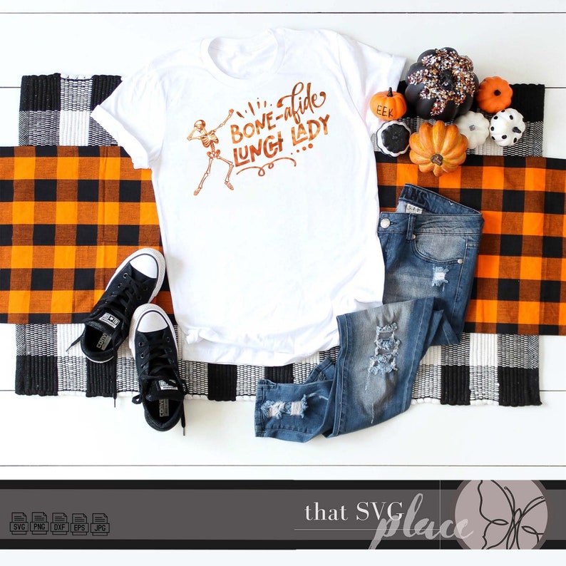 K&ouml;nnte beinhalten: Wei&szlig;es T-Shirt mit einem Skelett-Motiv und dem Text "Bone-afide Lunch Lady". Das Shirt ist auf einer karierten orange-schwarzen Decke mit einem Paar schwarzer Turnschuhe, einer blauen Jeans und kleinen orangefarbenen K&uuml;rbissen abgebildet.