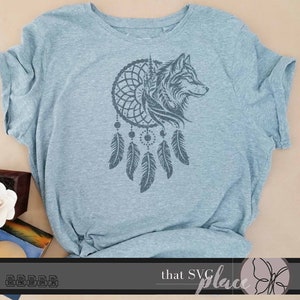 Op de afbeelding: Een grijs T-shirt met een zwart-witte afbeelding van een wolfskop in een droomvanger met hangende veren. De tekst "that SVG place" staat onderaan de afbeelding.