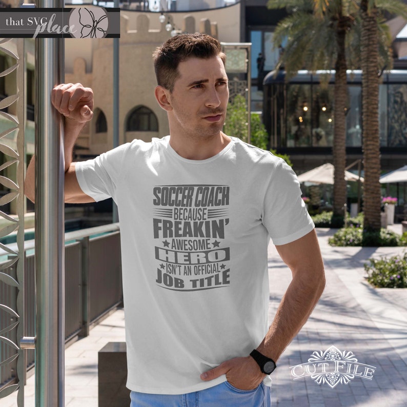 May include: White t-shirt with a gray graphic that reads "Soccer Coach Because Freakin' Awesome Hero Isn't An Official Job Title"