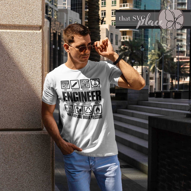 Op de afbeelding: Een wit T-shirt met een zwart grafisch ontwerp waarop "Engineer" staat en dat omringd is door een reeks waarschuwingssymbolen. De symbolen omvatten "Caution", "Hot", "Stop", "May Contain Nuts", "May Explode Under Pressure" en meer.