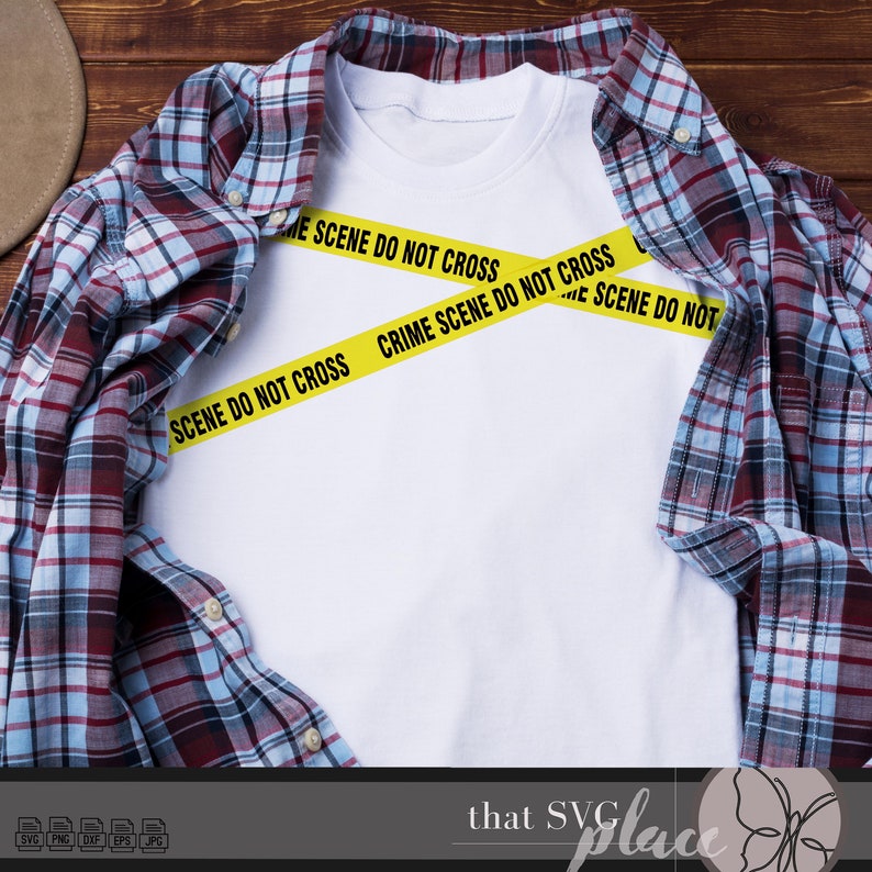 May include: A white t-shirt with yellow crime scene tape that says "Crime Scene Do Not Cross" across the front. The t-shirt is laying on a wooden surface with a plaid shirt on top of it.