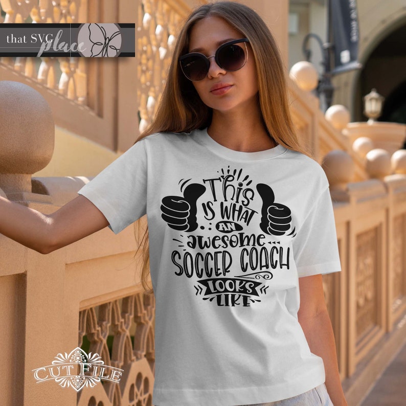 May include: White t-shirt with black text that reads "This is what an awesome soccer coach looks like" with two thumbs up on either side of the text.