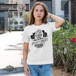 Op de afbeelding: Wit T-shirt met zwarte tekst die "Dit is hoe een geweldige receptionist eruitziet" zegt met twee duimen omhoog.