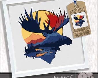 Puede incluir: Una obra de arte de alce 3D enmarcada. La obra presenta una silueta de alce en azul oscuro, con cuernos rojos, sobre un sol amarillo y un paisaje. Una etiqueta dice "3D MOOSE" y "3D SHADOW BOX".