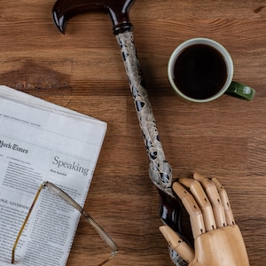 K&ouml;nnte beinhalten: Ein Gehstock aus Holz mit einem braunen und schwarzen Muster. Der Stock liegt auf einer Holzoberfl&auml;che mit einer Zeitung, einer Tasse Kaffee und einer Holzhand.