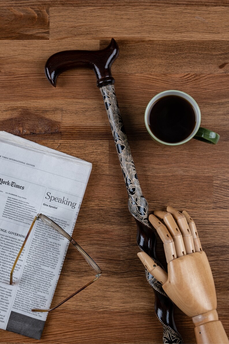 K&ouml;nnte beinhalten: Ein Gehstock aus Holz mit einem braunen und schwarzen Muster. Der Stock liegt auf einer Holzoberfl&auml;che mit einer Zeitung, einer Tasse Kaffee und einer Holzhand.