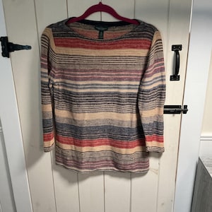 May include: A long-sleeved, striped sweater in shades of red, beige, blue, and purple hangs on a pink hanger. The sweater has a crew neck and a textured knit pattern. The background is a white wooden door.