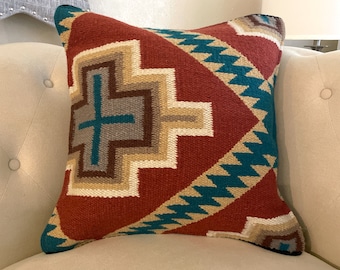 May include: Square decorative pillow with a geometric pattern in shades of red, teal, beige, and brown. The pillow features a central cross-like design and zig-zag borders, resting on a cream-colored upholstered surface.