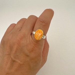 May include: A silver ring featuring an oval-shaped, orange spiny oyster shell. The shell has a natural, textured pattern. The ring is shown on a hand against a white background.