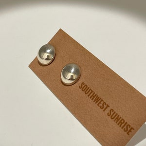 May include: A pair of shiny, silver-colored stud earrings with a smooth, rounded design. The earrings are displayed on a brown card with the text "SOUTHWEST SUNRISE" printed on it. The earrings reflect the surrounding environment.