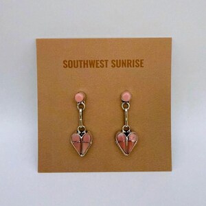 May include: A pair of silver-toned earrings with pink heart-shaped pendants. Each earring has a small pink circular stud at the top, connected by a chain to the heart-shaped pendant. The earrings are displayed on a brown card with the text "SOUTHWEST SUNRISE".