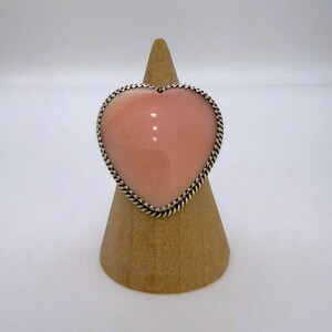 May include: A heart-shaped ring featuring a smooth, polished light pink stone set in a silver frame with a twisted rope-style border. The ring is displayed on a conical wooden stand, highlighting the contrast between the soft pink gemstone and the metallic silver setting.
