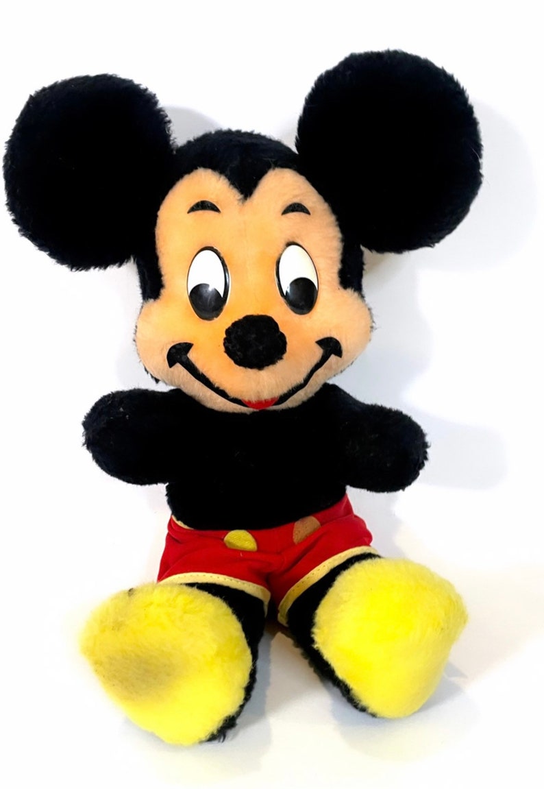 May include: A plush Mickey Mouse toy with black fur, large ears, and a red and yellow outfit. The toy has a smiling face with black eyes and a black nose.