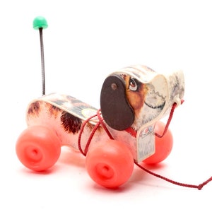Op de afbeelding: Een vintage trekdier in de vorm van een hond met een bruin-wit gevlekt lichaam, rode wielen en een groene antenne. De hond heeft een bruin oor en een rood touw dat aan zijn kop is bevestigd.