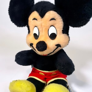 May include: A vintage plush Mickey Mouse toy with black fur, large yellow feet, and red shorts with yellow trim. The toy has a smiling face with black eyes and a black nose.