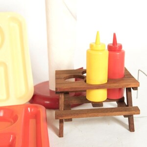 May include: A miniature wooden picnic table with two condiment bottles, one yellow and one red, on top. The table is made of brown wood and has a small bench on each side. The bottles are made of plastic and have a squeeze top.