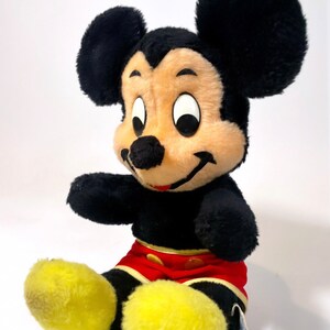 May include: A vintage plush Mickey Mouse toy with black fur, large ears, and a red and yellow outfit. The toy has a smiling face with black eyes and a black nose.