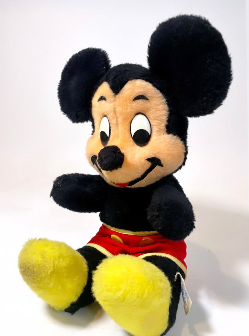 May include: A vintage plush Mickey Mouse toy with black fur, large ears, and a red and yellow outfit. The toy has a smiling face with black eyes and a black nose.