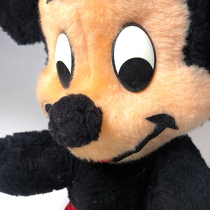 May include: A vintage plush Mickey Mouse toy with black fur, large white eyes, and a black nose. The toy is wearing red shorts with yellow trim.