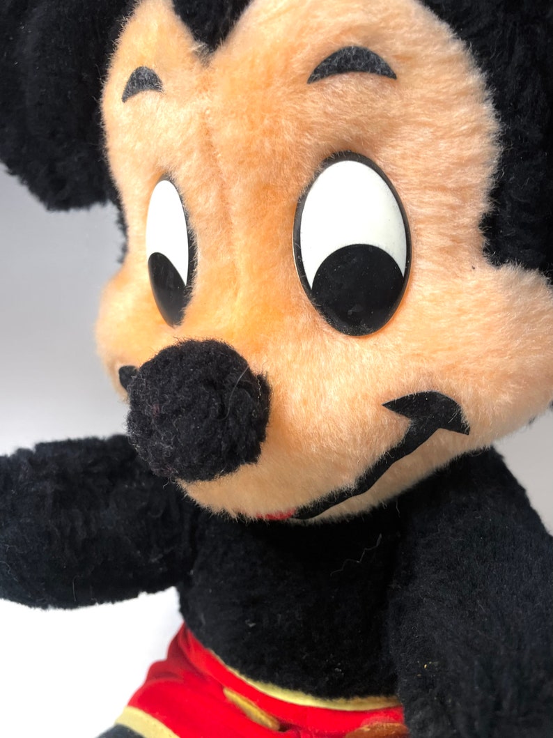 May include: A vintage plush Mickey Mouse toy with black fur, large white eyes, and a black nose. The toy is wearing red shorts with yellow trim.