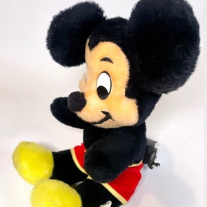 May include: A black and yellow plush Mickey Mouse toy sitting with a red and yellow shorts. The toy has a tag on its foot that says "Disney's Mickey Mouse".
