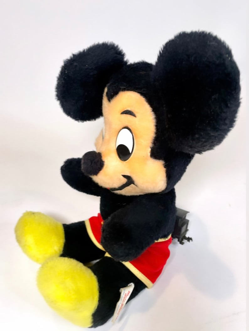 May include: A black and yellow plush Mickey Mouse toy sitting with a red and yellow shorts. The toy has a tag on its foot that says "Disney's Mickey Mouse".