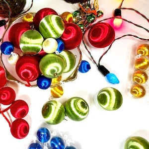 May include: Assortment of colorful satin Christmas ornaments in red, green, blue, and yellow. The ornaments are arranged in a bowl and on a white surface. Some of the ornaments have a striped pattern.