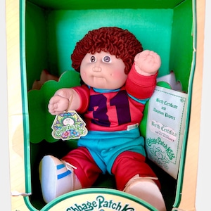 May include: A Cabbage Patch Kids doll with brown yarn hair, wearing a red and teal athletic jersey with the number 31 and matching pants. The doll is inside a green cardboard box with a birth certificate and adoption papers included.