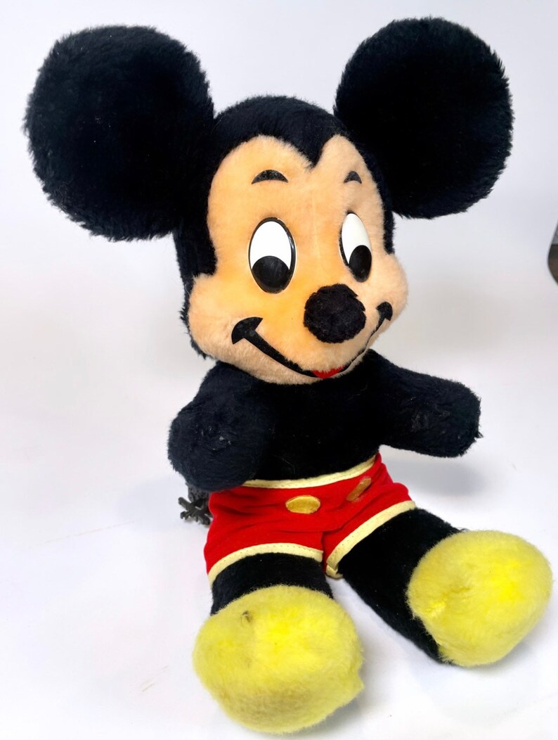 May include: A vintage plush Mickey Mouse toy with black fur, yellow shoes, and red shorts. The toy has a classic design with large ears and a friendly smile.