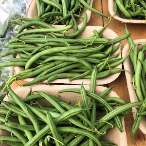 Op de afbeelding: Een close-up van een bos verse groene sperziebonen in een bruine papieren container. De bonen zijn in een stapel gerangschikt, met sommige die uit de container steken.