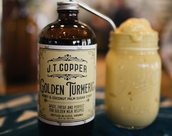 May include: A glass bottle of J.T. Copper Golden Turmeric Honey & Coconut Palm Sugar Syrup. The bottle is brown glass with a white label and black text. The label has a decorative design and the text reads "J.T. Copper Golden Turmeric Honey & Coconut Palm Sugar Syrup. Spicy, Fresh and Perfect for Golden Milk Recipes. Bottled in Floyd, Virginia. 16 fl oz (473 ml)".