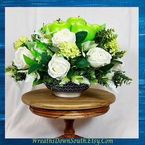 May include: A white ceramic bowl filled with green apples and white roses, surrounded by greenery. The bowl is sitting on a wooden pedestal.