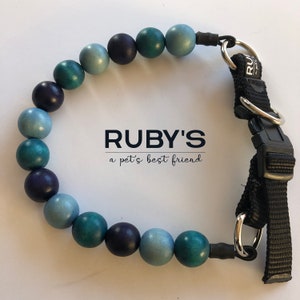 Pode incluir: Um colar de contas azuis e verdes para cães com uma alça preta ajustável e ferragens prateadas. O colar tem o texto "RUBY'S a pet's best friend" impresso nele.