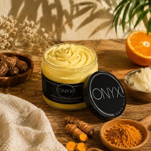 Op de afbeelding: Een open pot gele body butter met de tekst "ONYX Glow & Restore Body Butter" staat naast het zwarte deksel. Kurkumawortel, poeder en een halve sinaasappel liggen op een houten oppervlak.