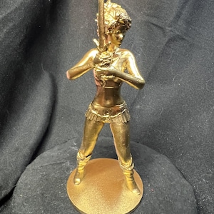 Puede incluir: Figura dorada de una mujer sosteniendo un bastón. La figura está de pie sobre una base redonda. La mujer lleva un top, una falda y botas. La figura está hecha de un material metálico brillante.