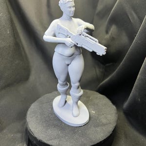 Puede incluir: Una figura gris de una persona sosteniendo un arma futurista. La figura está de pie sobre una base redonda, con detalles que incluyen botas y un corte de pelo corto. La estética general sugiere un tema de ciencia ficción o fantasía.