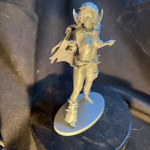 Op de afbeelding: Een grijze figuur van een vrouwelijke krijger, staand op een grijze ovale basis. De figuur draagt een cape, harnas en puntige accessoires. De figuur staat op een zwarte draaiende standaard.