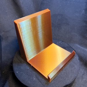 May include: A copper-colored phone stand with a rectangular base and a back support. The stand is made of a ridged material and has a slot to hold a phone. The stand is designed to hold a phone upright.