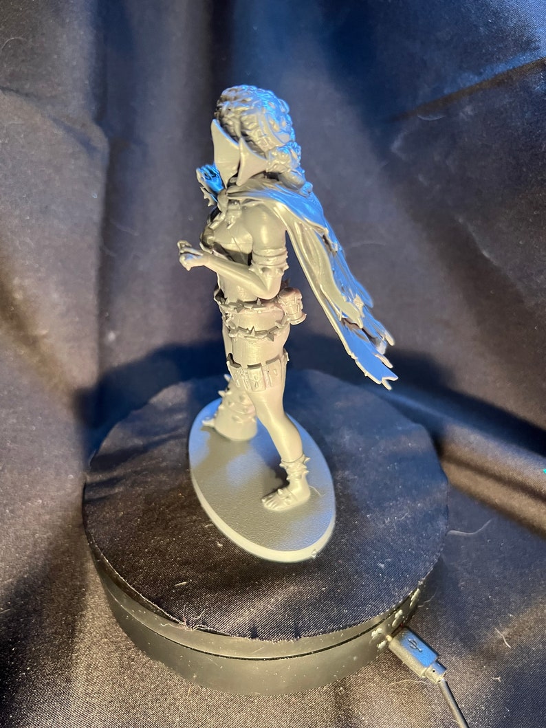 Op de afbeelding: Een grijze miniatuurfiguur van een krijgersvrouw, gekleed in een cape en harnas. De figuur staat op een grijze ovale basis. De krijger heeft een helm en houdt een klein voorwerp vast. Het beeldje is zeer gedetailleerd.