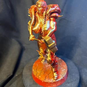 May include: A metallic gold figurine of a warrior woman, holding a hammer. The figure is posed on a circular base with a cracked, textured design. The armor has spiky shoulder pads and accents. The overall color scheme is gold and red.