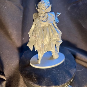 Op de afbeelding: Een zilverkleurig beeldje van een personage in een lange jas en cape, staand op een kleine, ovale basis. De figuur heeft gedetailleerd haar en gezichtskenmerken, met een dramatische pose. Het beeldje wordt getoond op een zwarte roterende standaard.