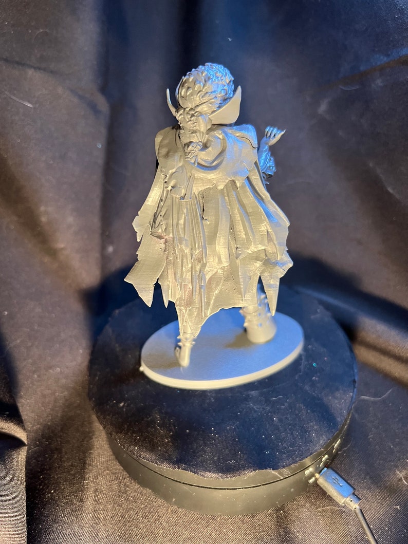Op de afbeelding: Een zilverkleurig beeldje van een personage in een lange jas en cape, staand op een kleine, ovale basis. De figuur heeft gedetailleerd haar en gezichtskenmerken, met een dramatische pose. Het beeldje wordt getoond op een zwarte roterende standaard.