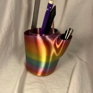 Puede incluir: Un organizador de escritorio impreso en 3D de color arcoíris con un bolígrafo, un lápiz y una pequeña herramienta en su interior.
