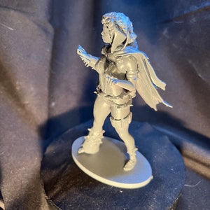 Op de afbeelding: Een grijze miniatuurfiguur van een persoon in een dynamische pose, met een cape, harnas en laarzen. De figuur staat op een grijze ovale basis. Het beeldje heeft gedetailleerde gelaatstrekken en kleding.