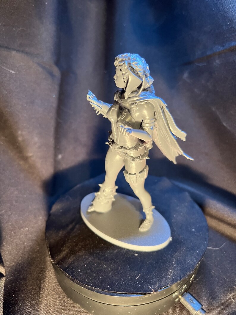 Op de afbeelding: Een grijze miniatuurfiguur van een persoon in een dynamische pose, met een cape, harnas en laarzen. De figuur staat op een grijze ovale basis. Het beeldje heeft gedetailleerde gelaatstrekken en kleding.
