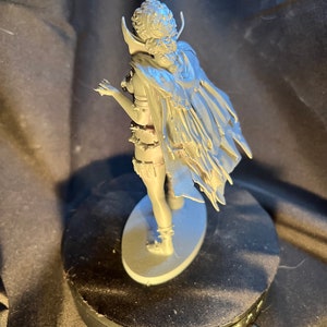 Op de afbeelding: Een grijze figuur van een personage met een cape en gedetailleerde kleding, staand op een ovale basis. De figuur poseert met &eacute;&eacute;n arm uitgestrekt. De figuur staat op een zwarte roterende displaystandaard.