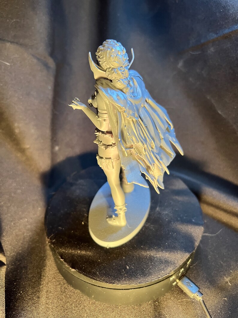 Op de afbeelding: Een grijze figuur van een personage met een cape en gedetailleerde kleding, staand op een ovale basis. De figuur poseert met &eacute;&eacute;n arm uitgestrekt. De figuur staat op een zwarte roterende displaystandaard.