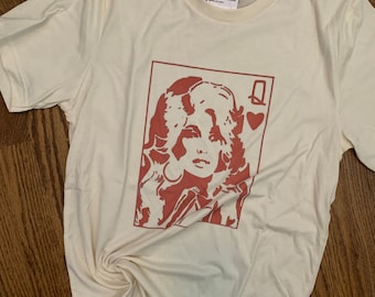 May include: A white t-shirt with a red silhouette of a woman with long blonde hair and a red heart with the letter Q on it. The text "Ramblin Rose" is on a tag attached to the shirt.