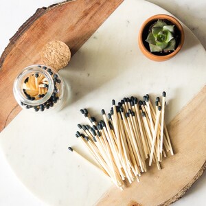 May include: A wooden round tray with a white marble inlay. The tray has a small potted succulent, a glass jar filled with matches, and a bundle of matches with black tips.