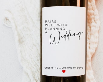 May include: A white wine bottle label with black text that reads "Pairs well with planning a Wedding. Cheers, to a lifetime of love." with a small red heart.
